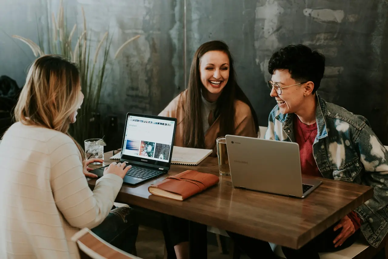 Três pessoas conversando sorridentes diante de um notebook em uma mesa de madeira